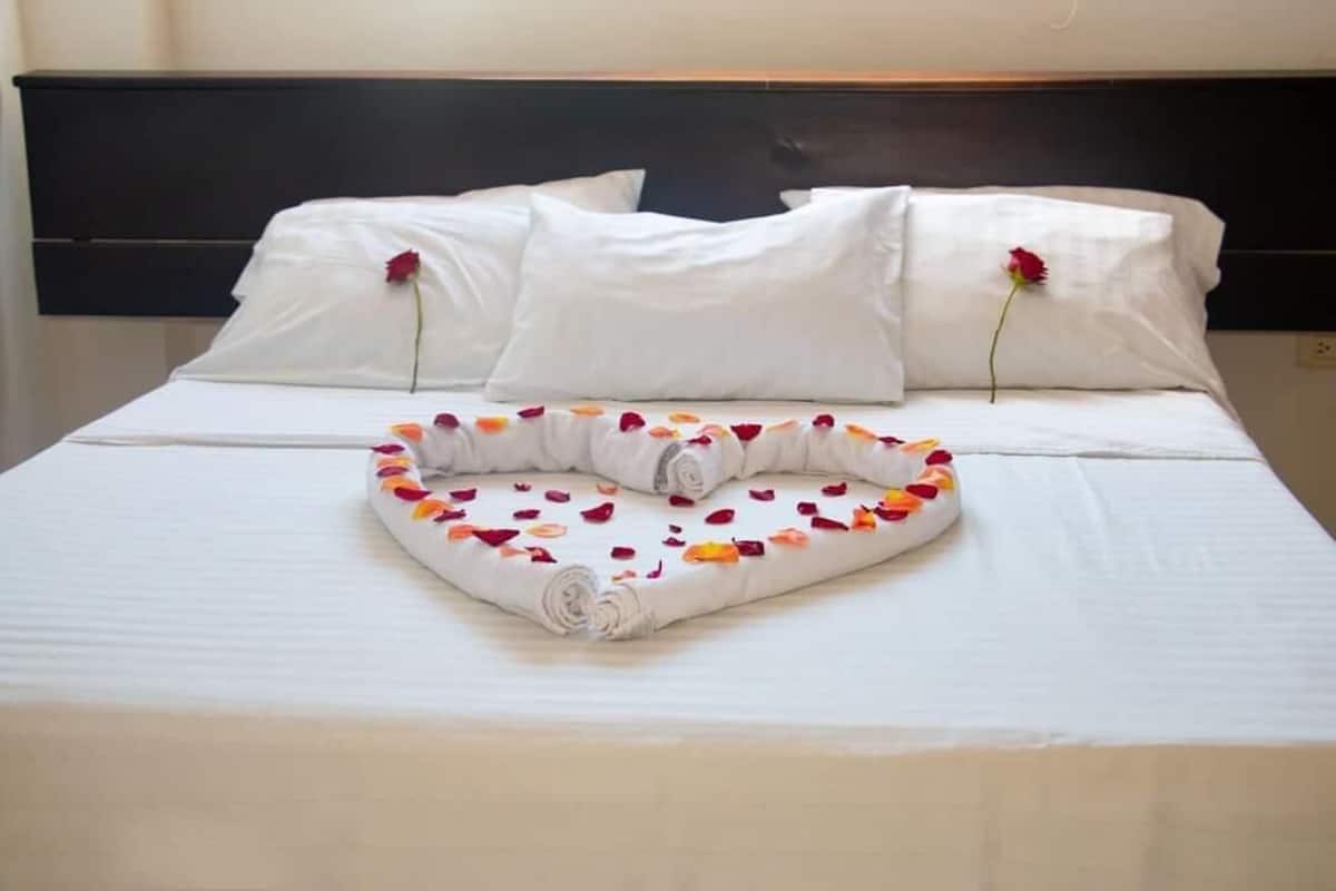 Una cama bien hecha con sábanas blancas, un arreglo de pétalos de rosa en forma de corazón y una toalla enrollada.