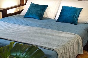 Una cama con colchón azul texturizado, dos almohadas, una manta de punto y una planta verde grande.