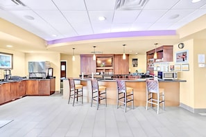 A modern kitchen with a central island, bar stools, a refrigerator, microwave, and a clock on the wall.