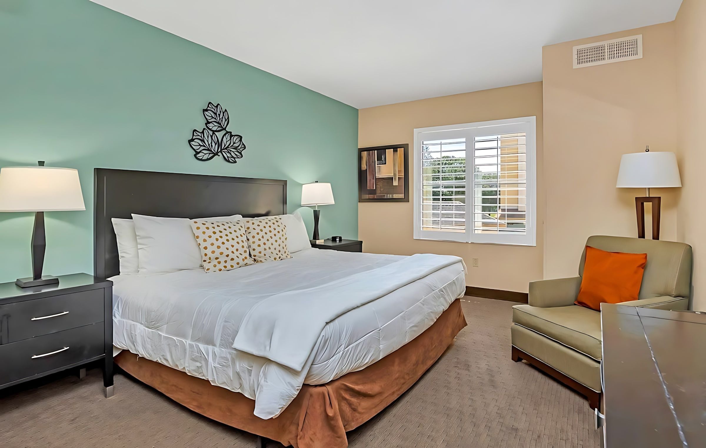 Une chambre d’hôtel comprenant un lit, deux tables de chevet avec des lampes, un canapé, une fenêtre avec des stores et un élément décoratif mural.