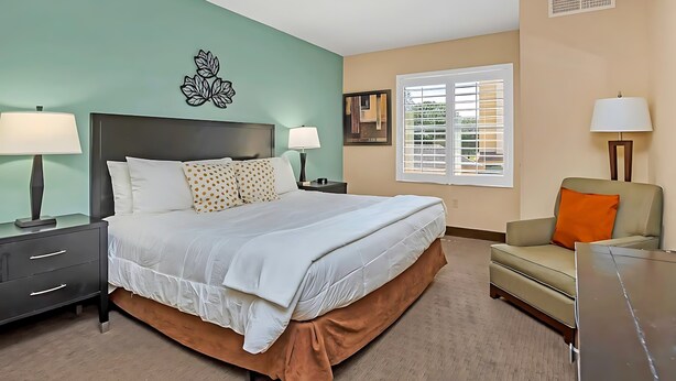 A hotel room with a bed, two bedside tables with lamps, a sofa, a window with blinds, and a wall decoration.