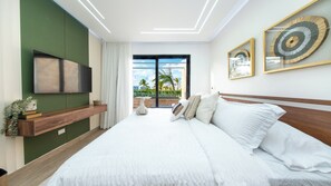 A modern bedroom with a large bed, a mounted TV, and a view of palm trees through the window. - Luxury 2-bedroom 2-bathroom condo with jacuzzi and huge pool in  Punta Cana (Punta Cana)