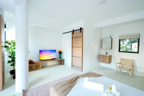 A modern hotel room with a flat-screen TV, a sliding door closet, a wooden bench, a rattan chair, a large window, and a potted plant.