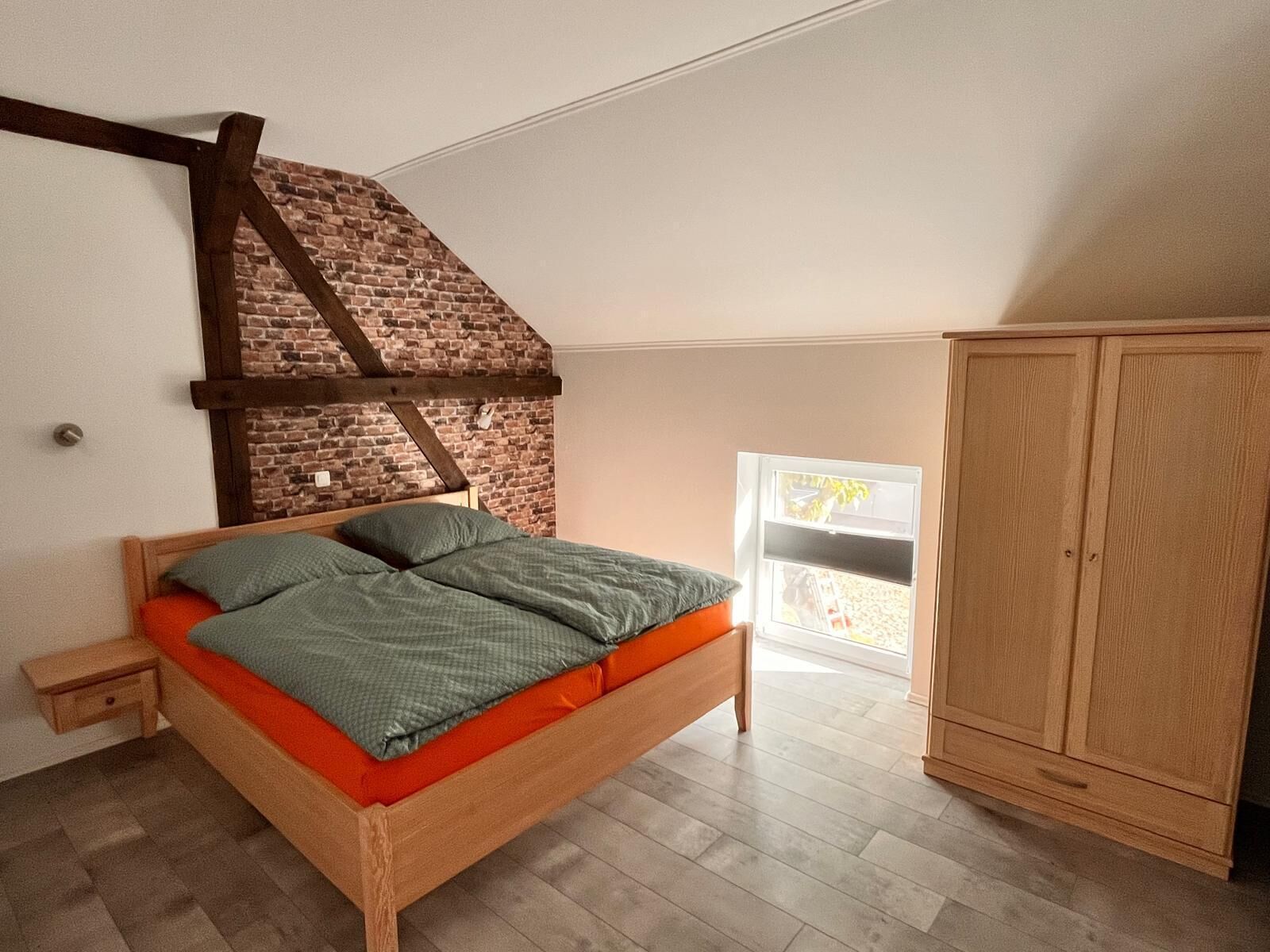 A bedroom with a sloped ceiling, a wooden bed, a wardrobe, and a view through a window.