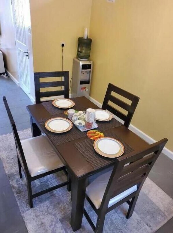 Comedor con mesa dispuesta para cuatro personas, sillas, dispensador de agua y una pequeña mesa con condimentos.
