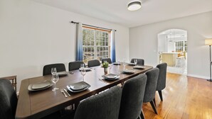 A dining area with a long table set for six, chairs, a window with curtains, and a view into the kitchen.