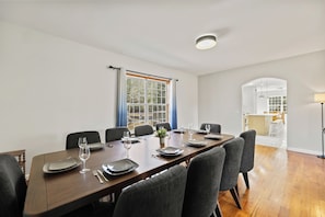 A dining area with a long table set for six, chairs, a window with curtains, and a view into the kitchen.