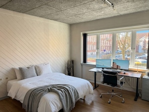 A bedroom with a bed, a desk with two monitors, a chair, and a view of the street through a window.