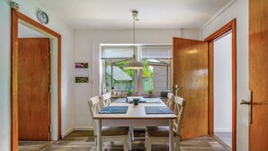 Un comedor con una mesa de madera, sillas blancas, una lámpara colgante y vistas de una casa verde a través del ventanal.