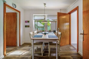 Un comedor con una mesa de madera, sillas blancas, una lámpara colgante y vistas de una casa verde a través del ventanal.