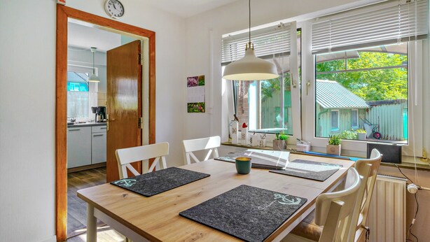 Un comedor con una mesa de madera, individuales negros, sillas blancas, una lámpara colgante y vistas a un jardín por la ventana.