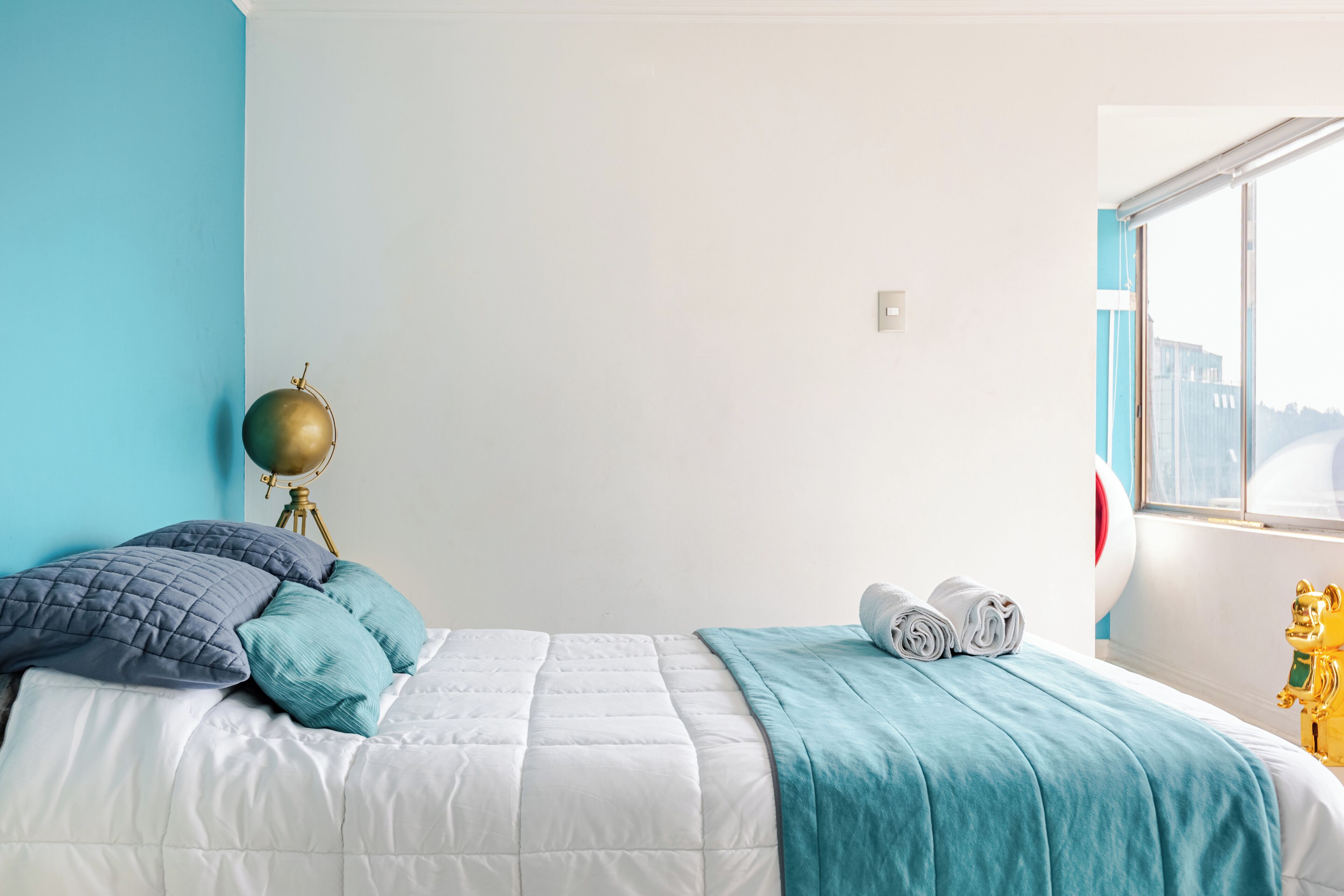 Un lit bien fait, recouvert d’une couvre-lit blanche et turquoise, un mur bleu et une fenêtre donnant sur des immeubles.