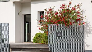 Entrada de um edifício com degraus que levam a uma porta, um portão de metal com uma placa e um arbusto com flores vermelhas.