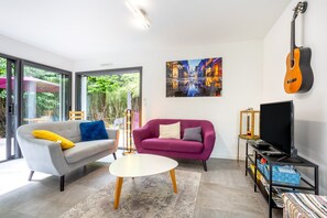 A modern living room with a grey sofa, a purple sofa, a white coffee table, a TV on a stand, and a guitar on the wall.