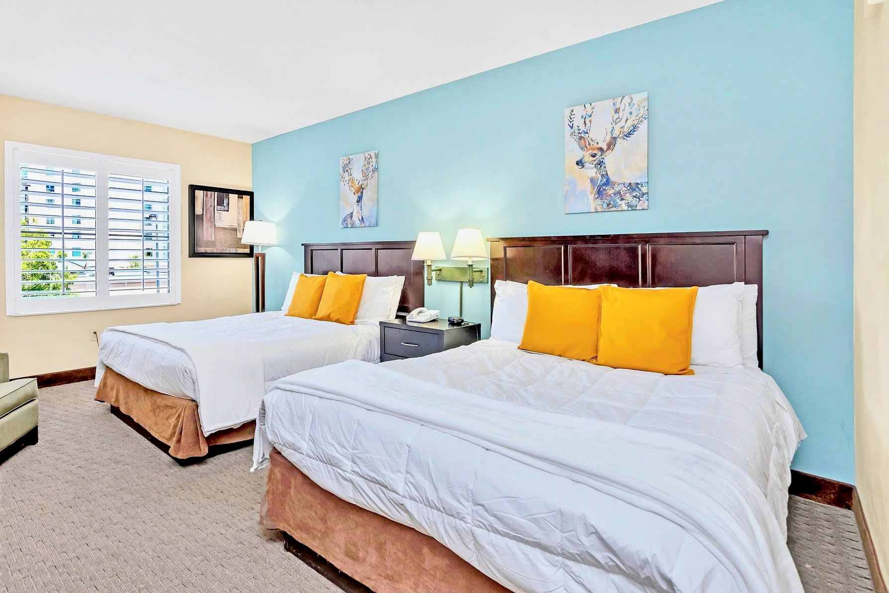 A hotel room with two beds, each with white bedding and yellow pillows. There are two wall-mounted lamps on either side of the beds. The walls are painted light blue, and there are two framed artworks above the beds.