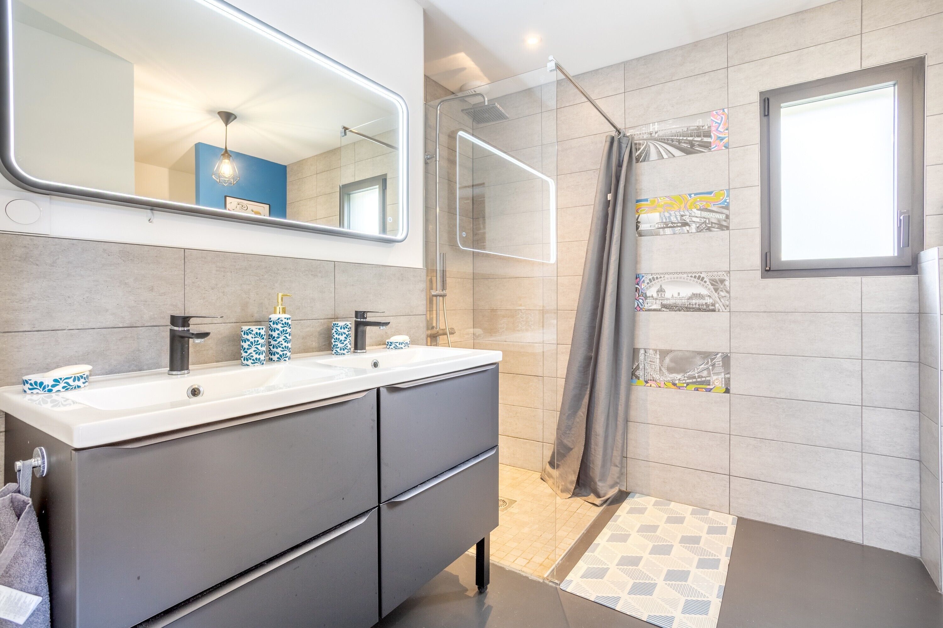 A modern bathroom with a dual sink vanity, a large mirror, a shower with a glass door, and tiled walls.