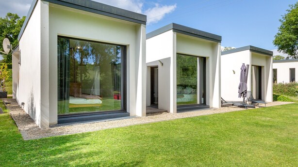 Une maison moderne à deux étages, dotée de grandes fenêtres en verre, d’un toit plat et d’une pelouse bien entretenue.