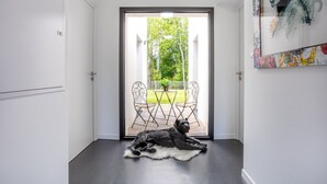 Une pièce où un chien noir est allongé sur un tapis blanc, une porte donnant sur un espace extérieur avec des chaises et une table, et un mur orné d’œuvres d’art abstraites.