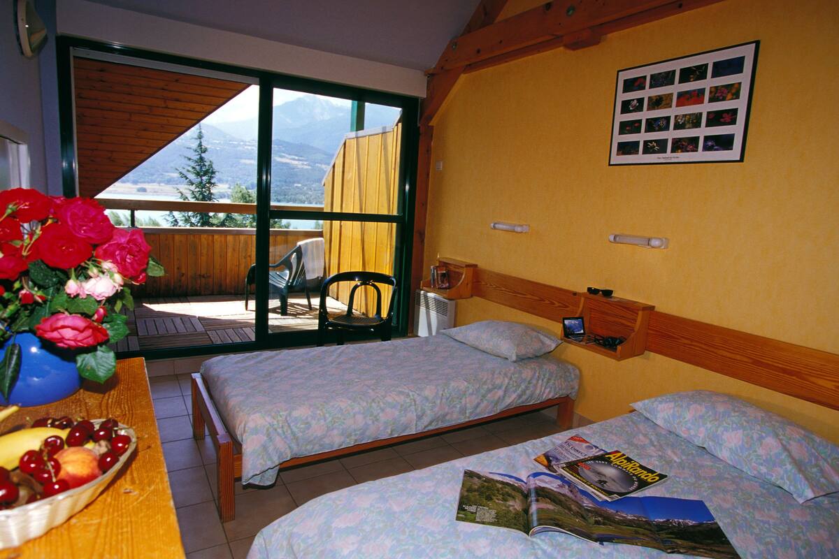 Ein Zimmer mit zwei Betten, einem Holztisch mit Obstschale, einem Stuhl und einem Fenster mit Bergblick.