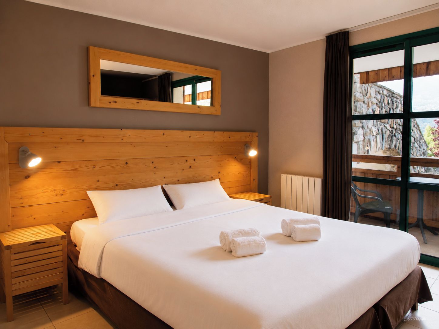A neatly made bed with white linens and two rolled towels, a wooden headboard, a bedside lamp, and a large window with a view of a stone wall.