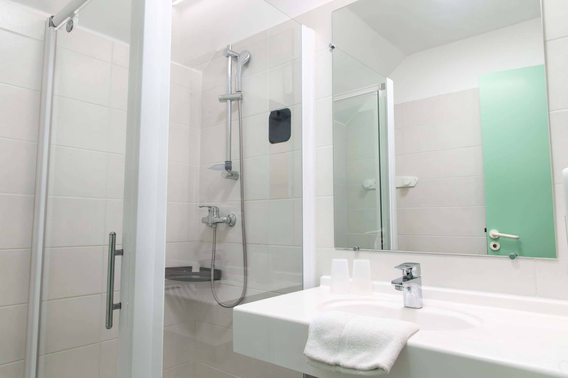 Un baño moderno con ducha envuelta en vidrio, un lavamanos blanco con una toalla y un espejo con detalles en verde.