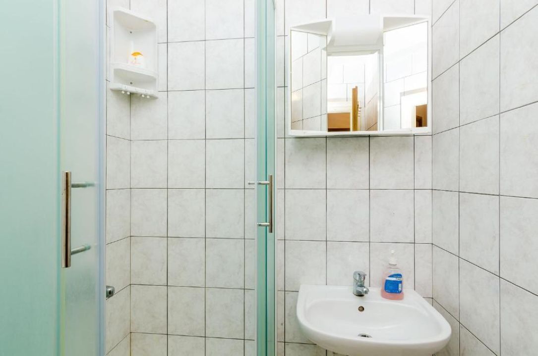 Een badkamer met witte betegelde wanden, een glazen douchedeur, een wastafel met zeepdispenser en een spiegel boven de wastafel.
