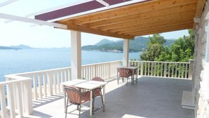 Ein überdachter Außenbereich mit Tisch und Stühlen, Blick auf das Meer und ein Steinhaus.