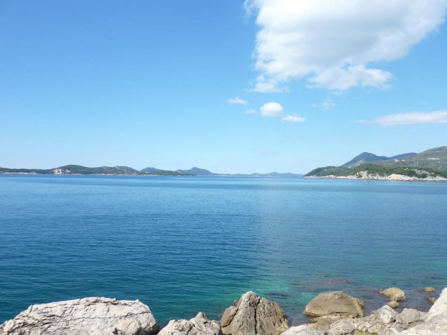A coastal landscape with calm water, rocky shoreline, and distant hills.