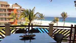 Un balcón con mesa y sillas, con vista a una playa con palmeras y una piscina.