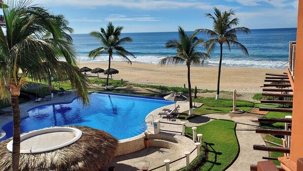 Una piscina con vista a la playa y al océano, rodeada de palmeras y sillones de descanso.