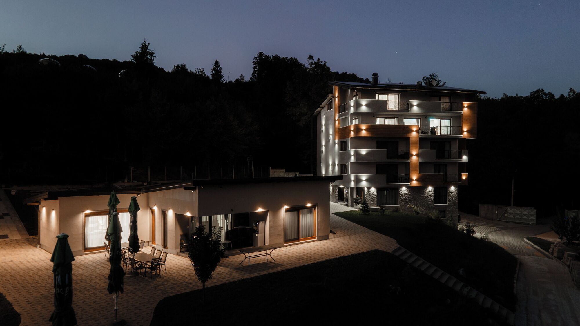 A modern multi-story building with illuminated balconies, a covered outdoor seating area, and a landscaped garden.