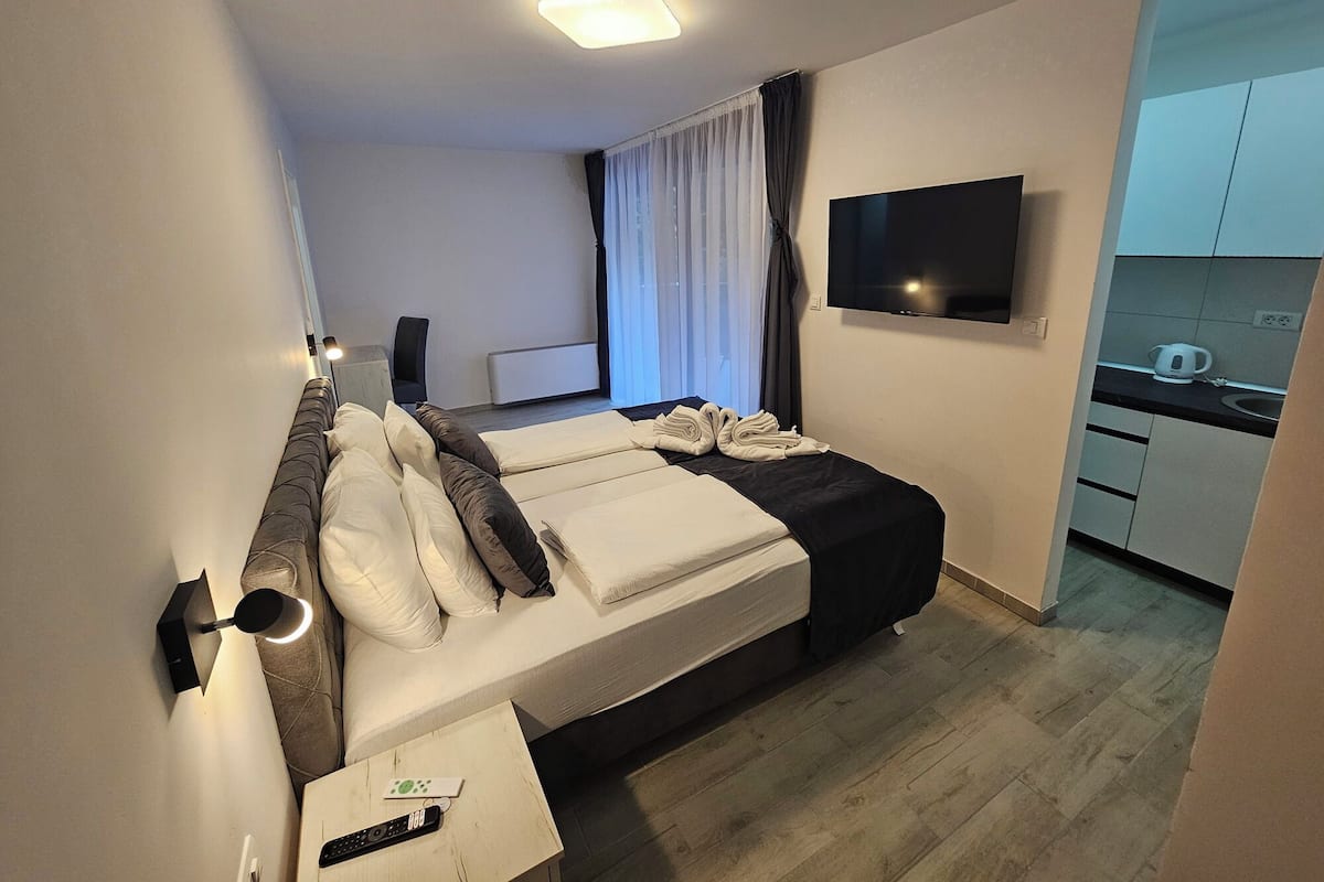 A hotel room with a bed, a TV, a small kitchenette, and a window with curtains.