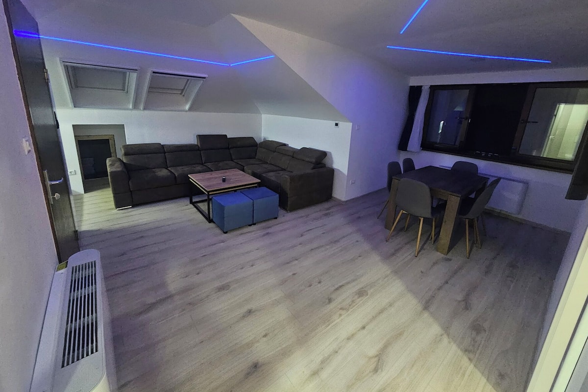 A modern loft-style room with a sectional sofa, a dining table with chairs, and a large window.