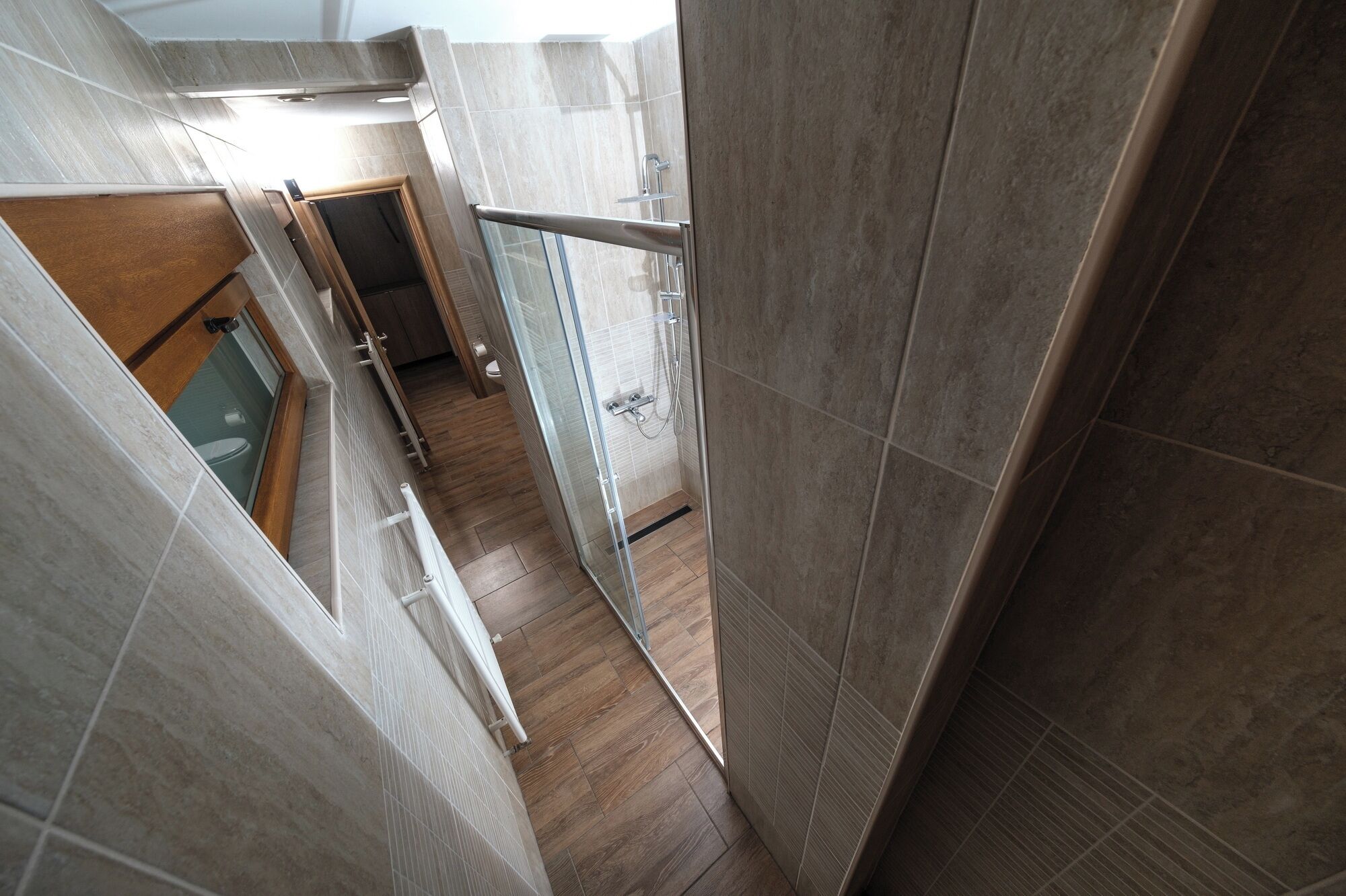 A narrow hallway with tiled walls and a wooden floor, leading to a glass shower area with a metal railing.