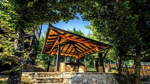 Um gazebo de madeira com base de pedra, rodeado por árvores e um céu claro.