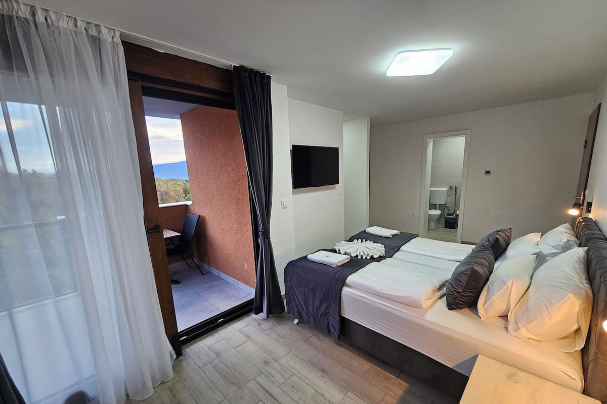 A hotel room with a bed, a TV, a balcony with a chair, and a view of the outdoors.