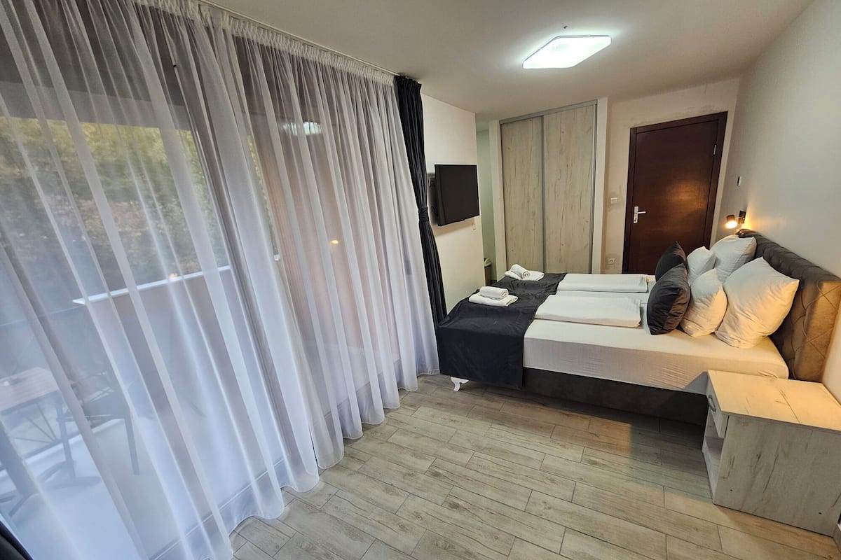 A hotel room with a bed, a TV, a wardrobe, and a window with sheer curtains.