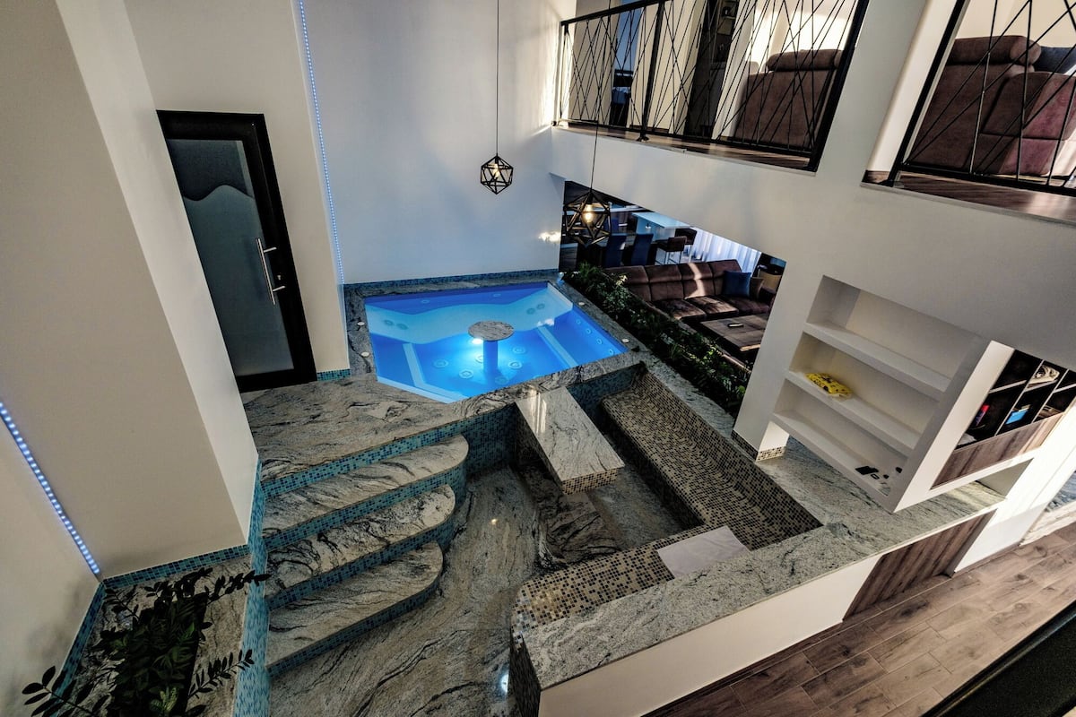 A multi-level indoor pool with a stone-tiled design, a hot tub, and a seating area with a view of the upper floor.