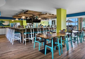 Ein Restaurant mit Barbereich, verschiedenen Sitzmöglichkeiten, darunter hohe Tische und Stühle, und Blick ins Freie.