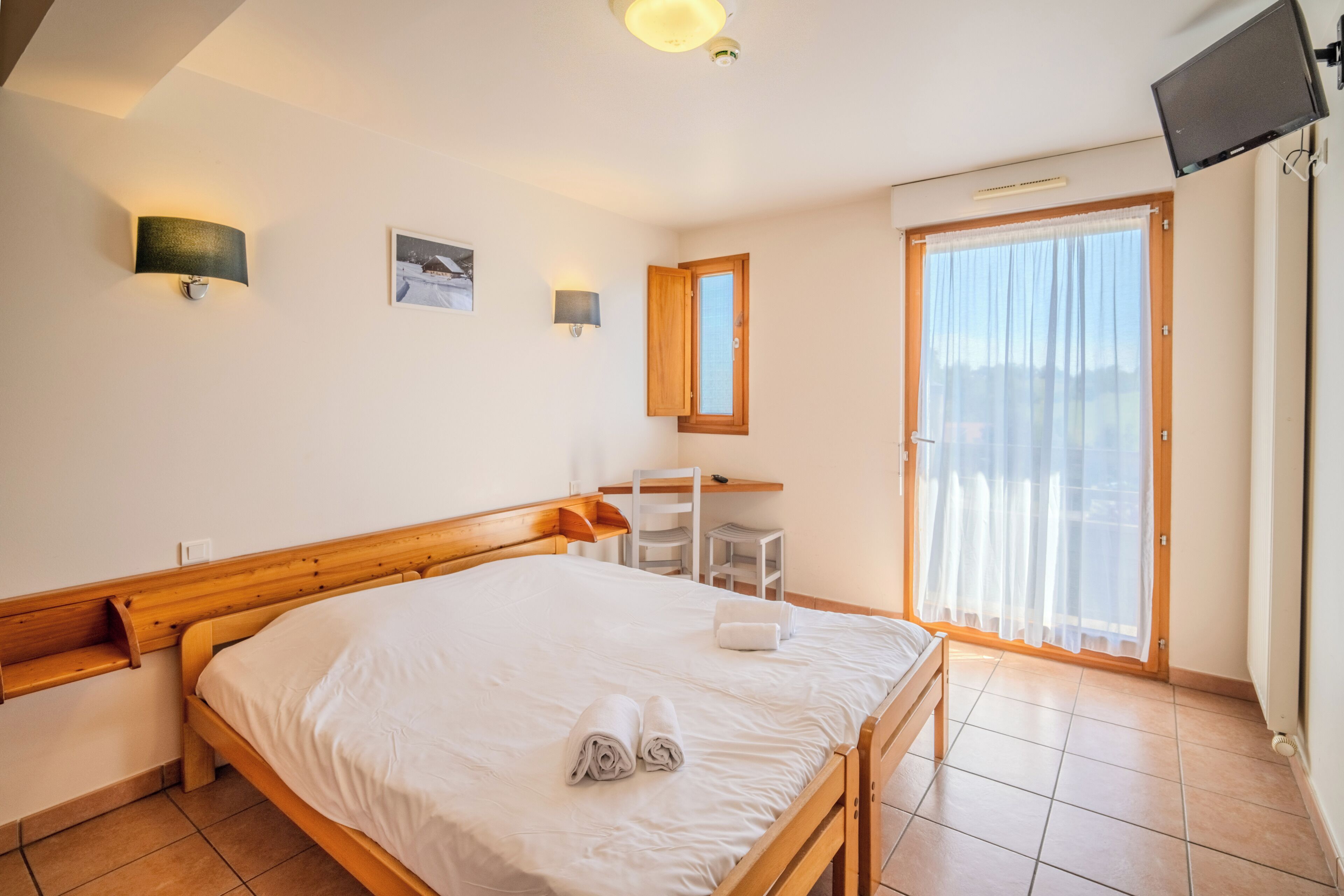 Ein Schlafzimmer mit einem hölzernen Bett, einem Schreibtisch, einem Stuhl, einem Fernseher und einer Schiebetür.