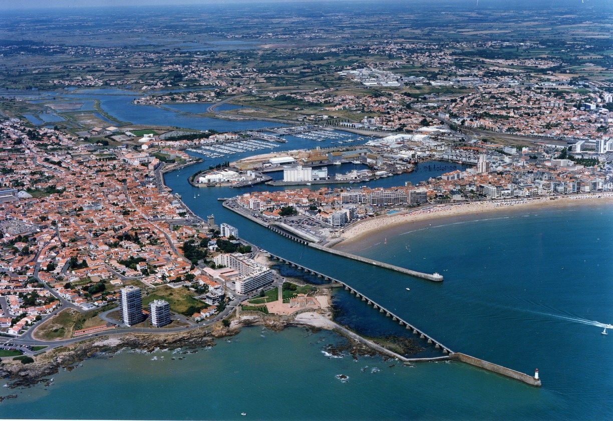 Vue aérienne d’une ville côtière avec un port de plaisance bien visible, une longue jetée et une zone de plage.