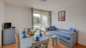 A room with a dining table, chairs, a sofa, a TV, and a window with curtains. - Proche du port | Balcon privé & vue sur le port (Les Sables d'Olonne)