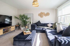 Ein modernes Wohnzimmer mit einem Ecksofa, einem Flachbildfernseher, einem Couchtisch, einer Topfpflanze und einem Wandbild.