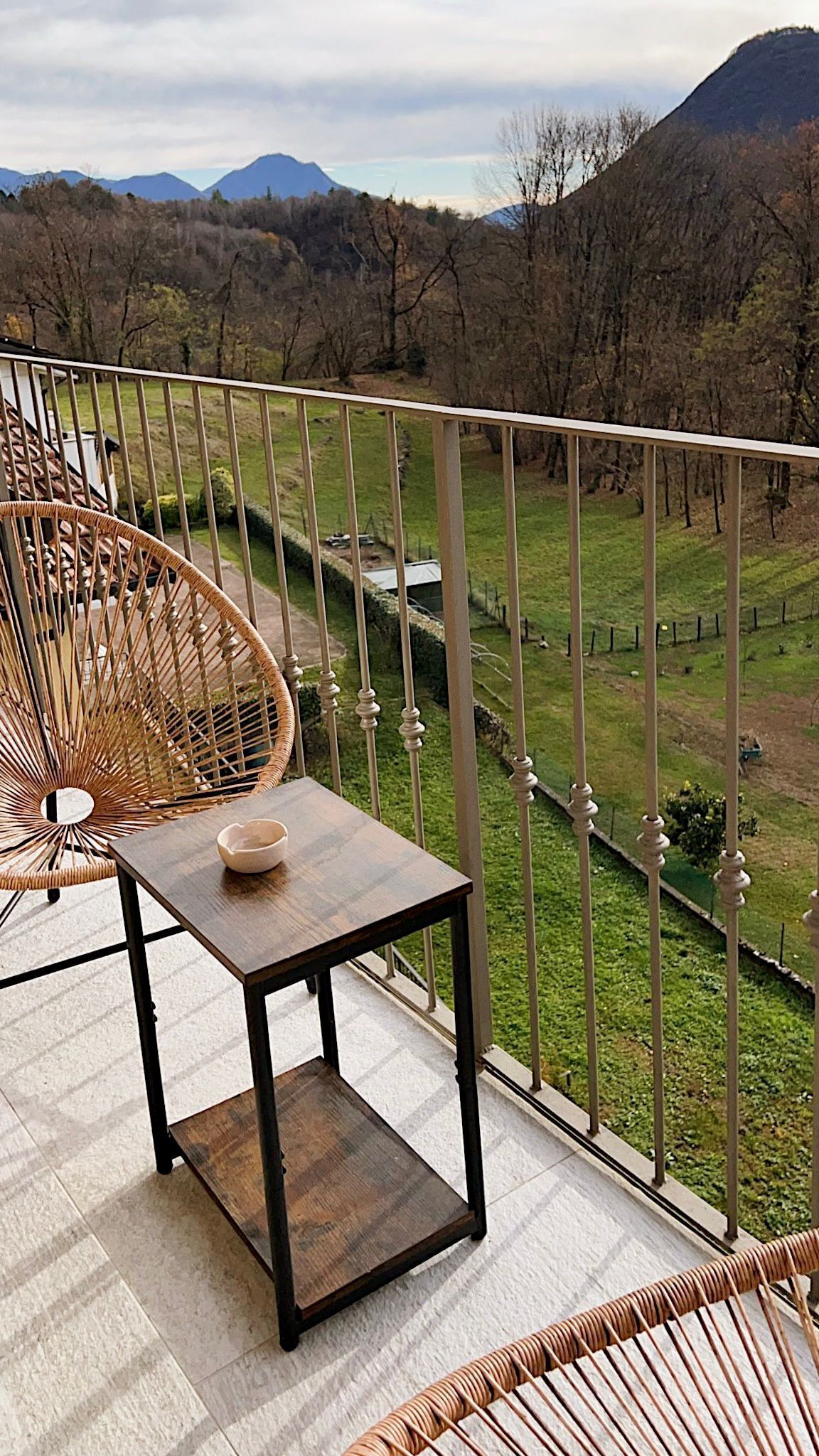 A balcony with a wicker chair, a small table, and a view of a green landscape with mountains in the distance.