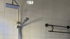 Une salle de bain avec des murs carrelés de carreaux blancs, une douche avec un pommeau de douche amovible, une barre à serviettes et un seau posé sur le sol.