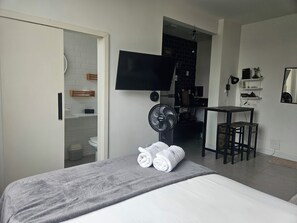 Habitación de hotel con cama, televisión, escritorio con silla, una mesita pequeña y baño a la vista a través de una puerta abierta.