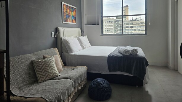 A bedroom with a bed, a sofa, a window with a city view, and an air conditioning unit. - Studio 5 minutes from Copacabana and Ipanema beaches (Rio de Janeiro)