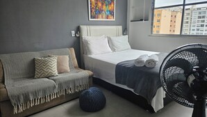 A modern room with a bed, a sofa, a fan, and a window with a city view. - Studio 5 minutes from Copacabana and Ipanema beaches (Rio de Janeiro)