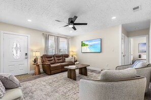 A living room with a brown leather sofa, a gray armchair, a wooden coffee table, a ceiling fan, and a large window with blinds. - Welcome to Tropical Breeze Oasis w/ heated pool and relaxing back yard & patio (Sarasota)