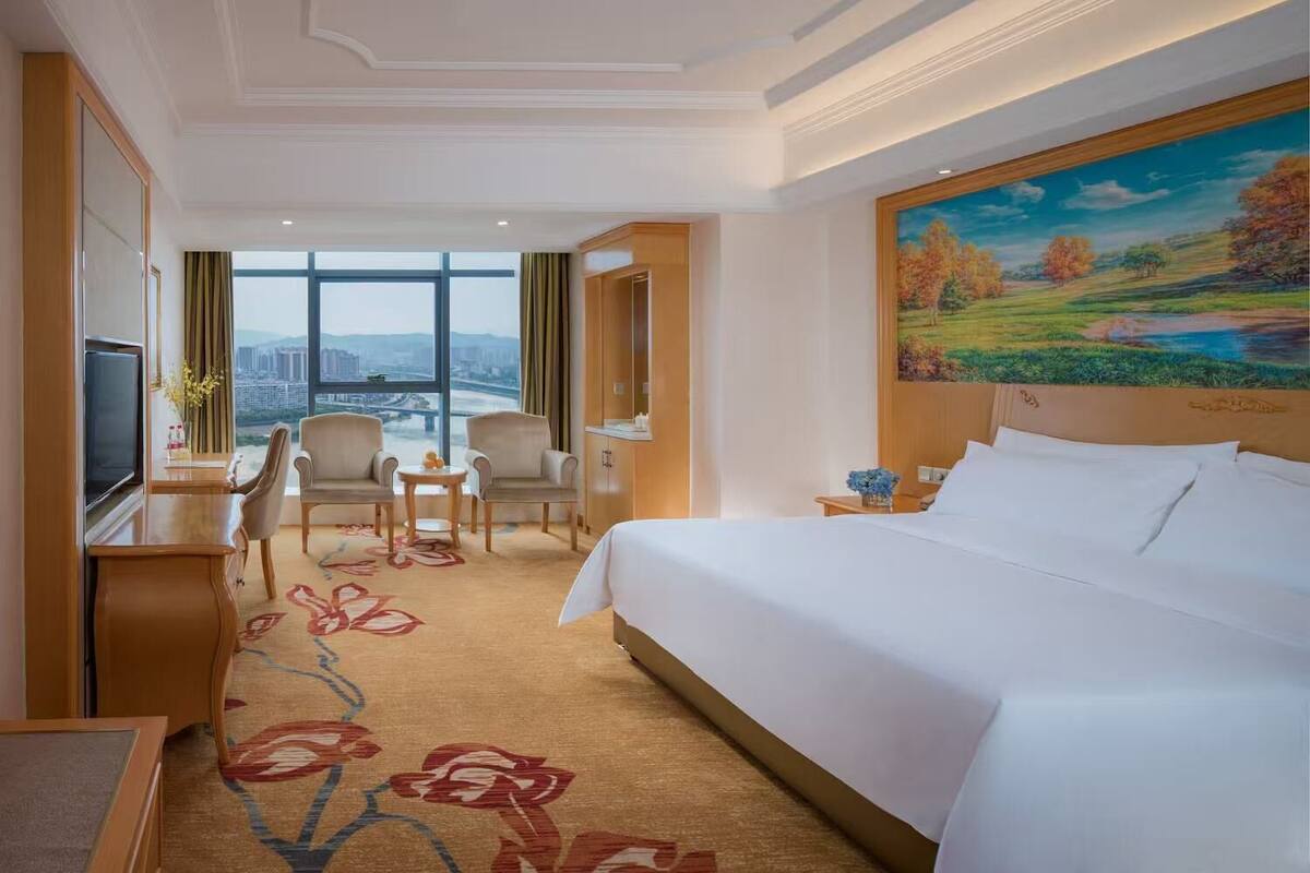 A hotel room with a bed, a desk with a TV, two chairs, a small table, and a painting on the wall.